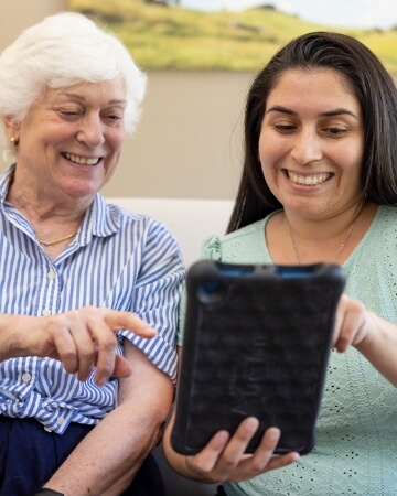 An elderly woman with a bright smile enjoys looking at a tablet with a young woman, illustrating a friendly interaction at Carlton Senior Living.