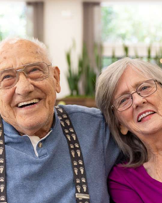 A cheerful elderly couple smiles while seated together in a bright, cozy space at Carlton Senior Living.