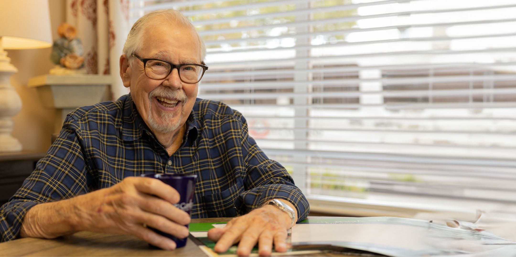 An elderly man in a plaid shirt smiling while holding a coffee mug at a table, symbolizing a comfortable and secure retirement.