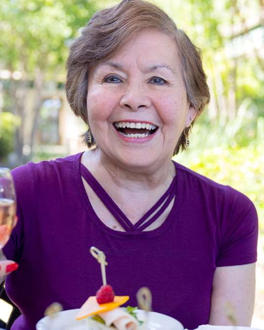 An elderly woman holding a beverage, laughing joyfully outdoors, surrounded by greenery at a Carlton Senior Living Independent Living community.