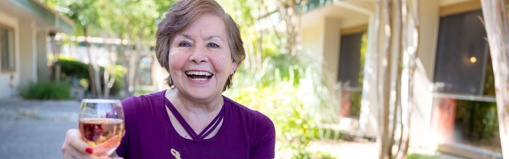 An elderly woman holding a beverage, laughing joyfully outdoors, surrounded by greenery at a Carlton Senior Living Independent Living community.