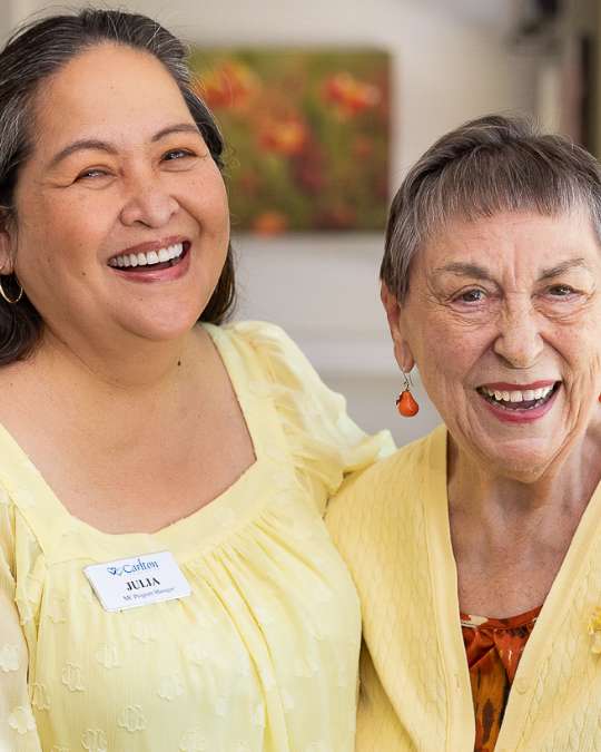 A Carlton Senior Living team member smiling alongside a cheerful Memory Care resident, both dressed in yellow, inside a bright and welcoming environment.