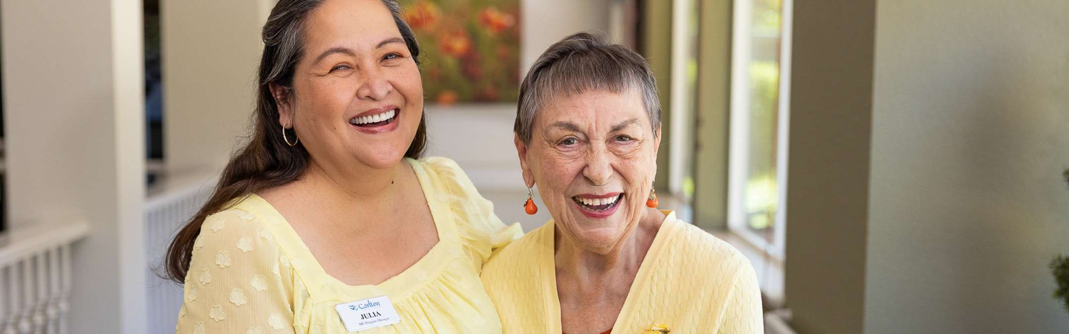 A Carlton Senior Living team member smiling alongside a cheerful Memory Care resident, both dressed in yellow, inside a bright and welcoming environment.