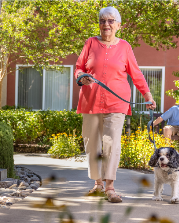 An elderly woman walking her dog along a pathway in a sunny courtyard, symbolizing an active and fulfilling lifestyle in senior living.