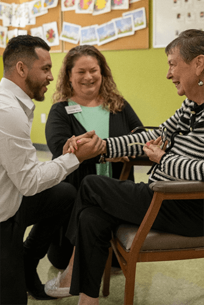 Two Carlton Senior Living team members engaging with a senior resident, holding hands and sharing a moment of connection, highlighting the Positive Approach to Care.