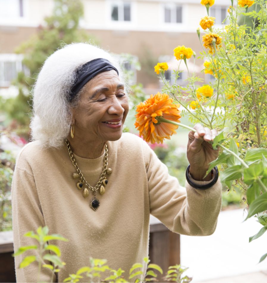 An elderly woman smiling as she smells a bright orange flower in a garden, representing a peaceful and joyful retirement.