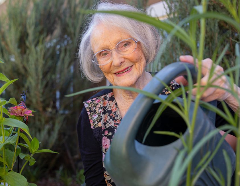 An elderly woman with glasses watering plants in a garden, highlighting a fulfilling lifestyle in retirement.