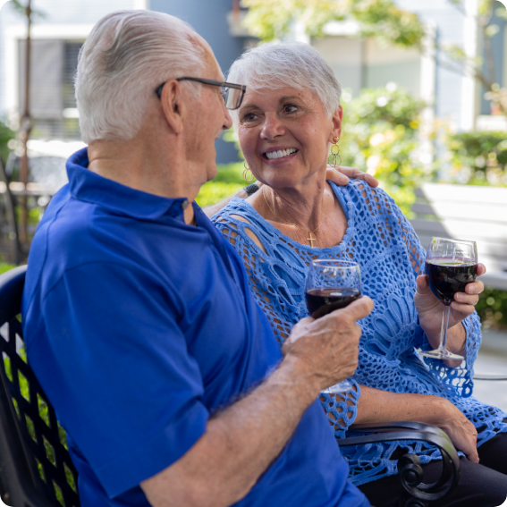 A senior couple sitting outdoors, enjoying a glass of wine together and smiling warmly at each other.